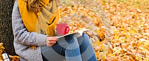 Banner. Young woman drinks hot tea or coffee from a mug and reads a book in the park while sitting under a tree. Autumn mood.