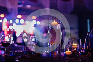 Banquet table decorated with burning candles in glass vases in restaurant hall. In the background party with silhouettes