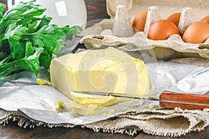 A bar of butter is cut into pieces on a wooden board with a knife, surrounded by milk, eggs and parsley on a brown table. Ingredie