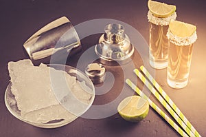 bar concept: Tequila shots, lime, ice, straws and shaker/Tequila shots, lime, ice, straws and shaker on a dark background. Toned.