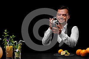 A bartender wipes a shaker at the bar counter, lemon, lime, oranges, cocktails on a black background.