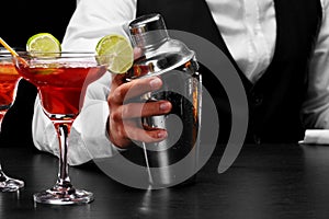 A shaker on a bar counter, a margarita glass full of cocktail, a hand of a bartender on a black blurred background.