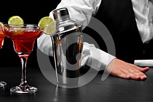 A shaker on a bar counter, a margarita glass full of cocktail, a hand of a bartender on a black blurred background.