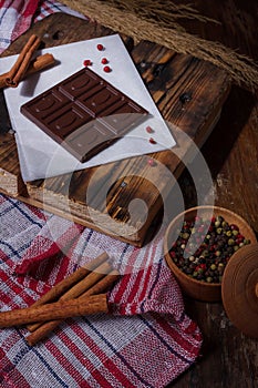 Bar of dark bitter chocolate with chilly pepper, cinnamon, red pea pepper flavour on white paper, wooden board background. Unusual
