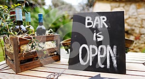 `Bar is open` sign and wooden crate full of wine bottles decorated with olive branches on a table. Festive party, weddin