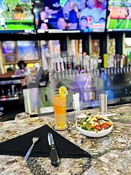 Bar table with silverware on cloth napkins, tall pilsner beer glass, healthy salad bowl and blurry beer taps, wall mount big