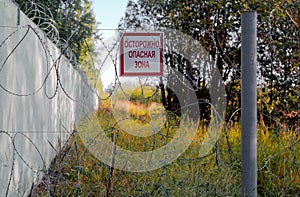 On a barbed wire fence near a high concrete wall there is a sign with the inscription in Russian