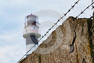 barbed wire and stone fence, behind them a lighthouse. concept of slavery, dictatorship and totalitarianism