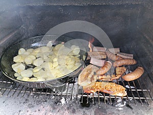 Barbequing pork meat and sausages with dollar chips potatoes close up