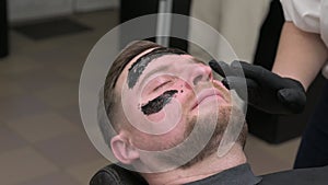 Barber applies a black moisturizing mask to a man's face while shaving. A dermatologist applies a black mask to the