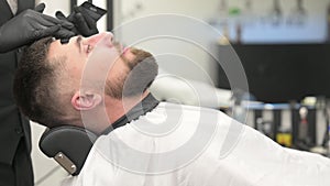 Barber applies a black moisturizing mask to a man's face while shaving. A dermatologist applies a black mask to the