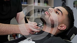 A barber cuts the beard of a man in a barbershop with an electric razor.