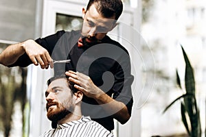 Barber dressed in a black shirt with a red bow tie makes a stylish hairstyle to young man in a barbershop