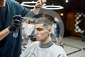 Barber fixing haircut of his client with hair spray while working in the barbershop. Young man visiting barbershop