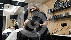 A barber girl in a protective mask shaves a young bearded Caucasian man with a straight razor. A young girl in a barber