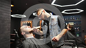 Barber in glasses cuts man`s beard with a trimmer in barbershop. Hairstyling and hair cutting in salon. Grooming the