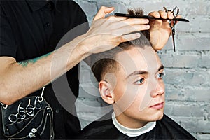 Barber hair styling of young guy in the barbershop on brick wall background, hairdresser makes hairstyle for a young man.