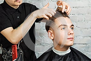Barber hair styling of young guy in the barbershop on brick wall background, hairdresser makes hairstyle for a young man.
