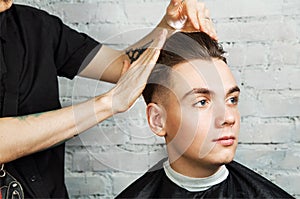 Barber hair styling of young guy in the barbershop on brick wall background, hairdresser makes hairstyle for a young man.