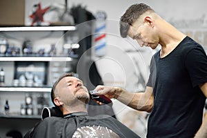 Barber master is shaving of handsome mature bearded man in salon. Hair artist making hairstyle for person in barbershop. Services