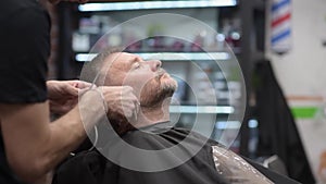 Barber master shaving handsome mature bearded man using electric shaver in salon. Hair artist making beard style for person in mal
