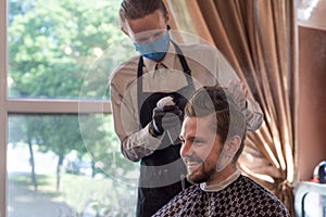 A barber in a protective mask in a barbershop is cutting a young bearded guy. quarantine haircut, barbershop worker in a