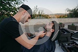 Barber shaves beard of client man on chair an open air