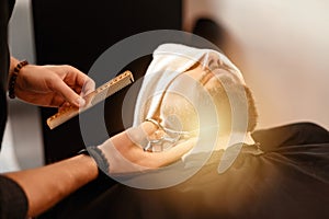 A barber stylist trims the beard of a Caucasian man, whose face is covered with towel, with scissors