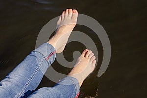 Bare feet of a girl in jeans over dark water on a summer sunny day. Horizontal close-up photo