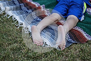 Bare feet and legs of a young person lying stomach down on a woven blanket on the grass at an outdoor concert