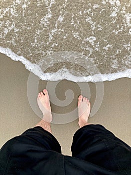Bare feet of a man standing on sand by the sea while a wave comes in. Concept of vacation, relaxation, freedom or refreshment. POV
