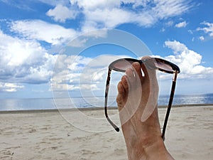 Bare feet raised up with colored sunglasses on the beach. Beautiful sea surf and blue sunny sky in background