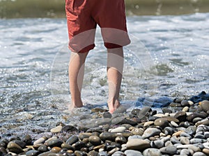 Bare feet of an unspecified boy in the water on the coast with stone beach
