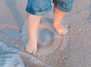 Bare feet walking at sandy beach near the sea. Little baby in blue jeans shorts going to touch the sea at sunset. Wave