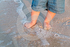 Bare feet walking at sandy beach near the sea. Little baby in blue jeans shorts going to touch the sea at sunset. Wave