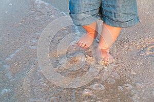 Bare feet walking at sandy beach near the sea. Little baby in blue jeans shorts going to touch the sea at sunset. Wave