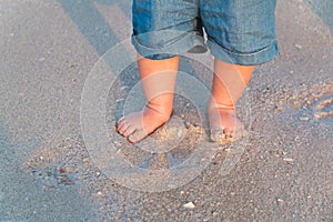 Bare feet walking at sandy beach near the sea. Little baby in blue jeans shorts going to touch the sea at sunset. Wave