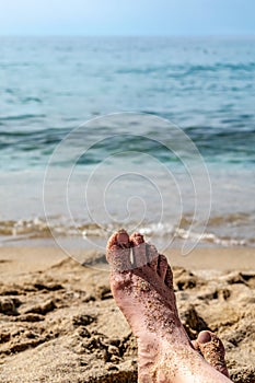 Bare female foot soiled with sand against the background of blue sea water, vertical. Beach summer background with copy space.