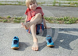 barefoot boy sits on the asphalt and children\'s blue sneakers stand nearby