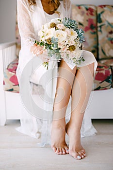 Barefoot bride in a lace robe sits on a sofa in a hotel room and holds a wedding bouquet in her hands, close-up