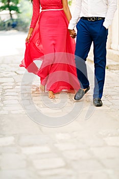 Barefoot bride in a long bright pink dress and the groom walking along a cobbled road and tenderly hold hands