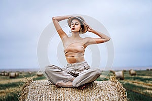 Barefoot brunette in linen pants and bare shoulders sitting on a hay bales in warm autumn day. Behind her is a wheat field