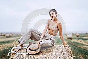 Barefoot brunette in linen pants and bare shoulders sitting on a hay bales in warm autumn day. Woman looking at camera. Behind her
