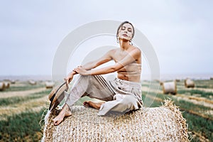Barefoot brunette in linen pants and bare shoulders sitting on a hay bales in warm autumn day. Woman looking at camera. Behind her