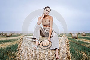 Barefoot brunette in linen pants and bare shoulders sitting on a hay bales in warm autumn day. Woman looking at camera. Behind her