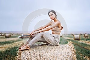 Barefoot brunette in linen pants and bare shoulders sitting on a hay bales in warm autumn day. Woman looking at camera. Behind her