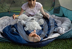 barefoot child sits in a tent, covered with a sleeping bag, and plays with favorite teddy bear