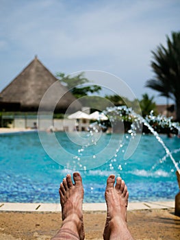 Barefoot feet of a man taking a sun bath in a pool with fountains nd a hut on the background photo
