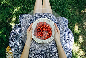 Barefoot girl is sitting on the grass in blue vintage dress. Woman is holding plate with home strawberries. Rustic summer fruit