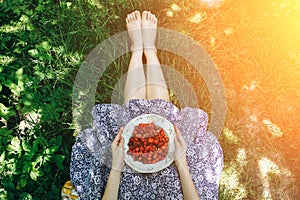 Barefoot girl is sitting on the grass in blue vintage dress. Woman is holding plate with home strawberries. Rustic summer fruit